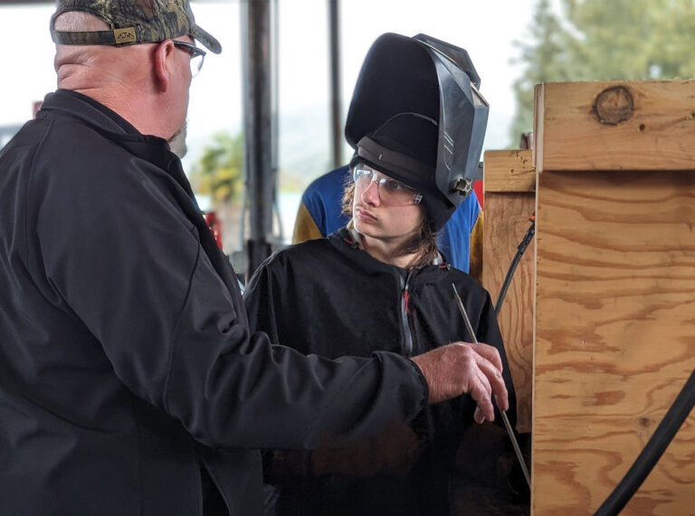 A young member of the community in a welding mask learns from a Con-Vey leader